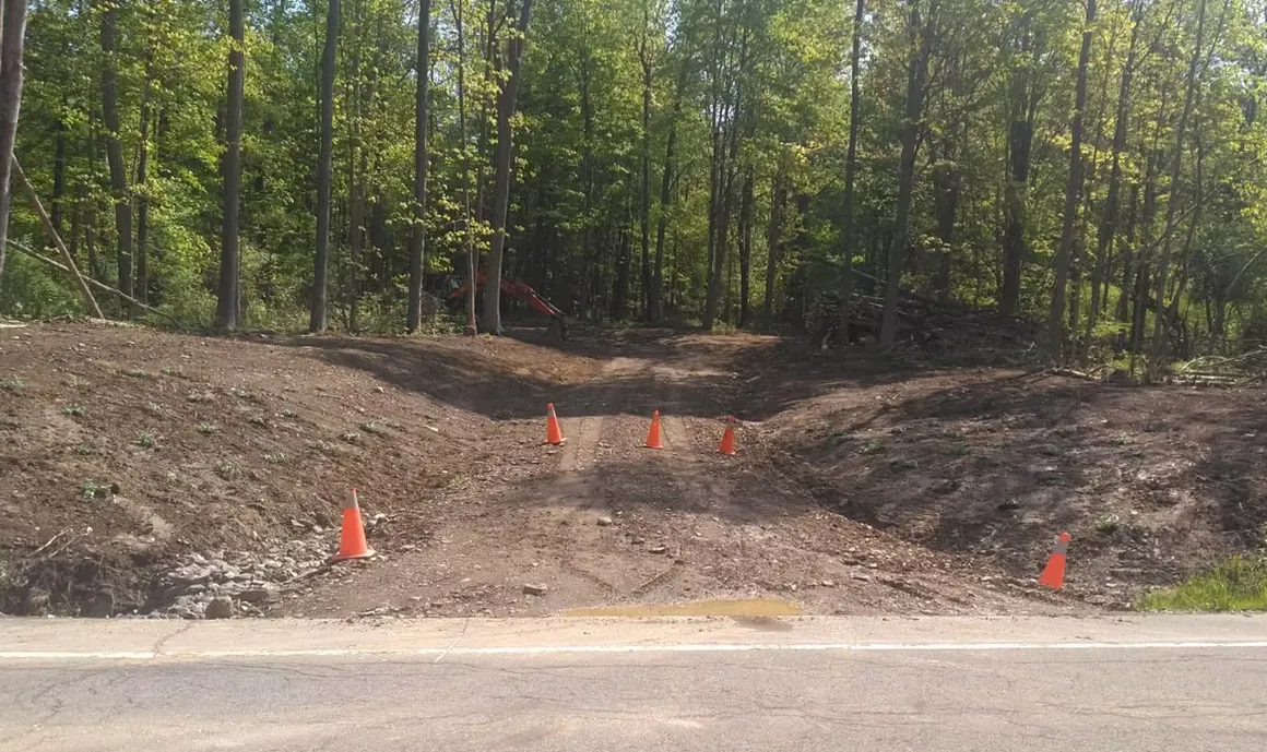 Landmark Enterprises site excavation with excavator and road-edge driveway cut marked with safety cones in Dewittville, NY